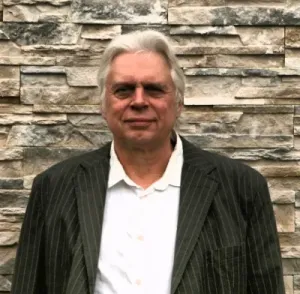 Brustbild Jürgen Langowski vor einer Mauer, weiße Haare, weißes Hemd, graues Jackett.