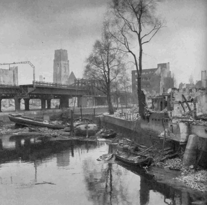 Vordergrund: Trümmer an einem Kanal, im Hintergrund eine zerstörte Brücke