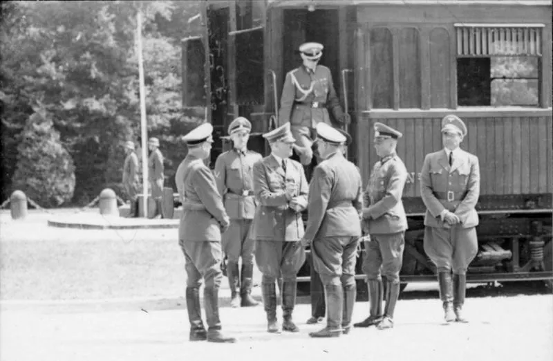 Im Hintergrund ein Eisenbahnwaggon, aus dem ein Uniformierter des NS-Militärs steigt. Davor stehen Hitler und weitere Uniformierte.
