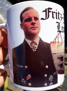 Ein Gedenk-Kaffeepott, mit Brustbild eines Offiziers mit Ritterkreuz, rechts oben in Frakturschrift der Name. Rechts im Hintergrund ein nicht erkennbares Bauwerk.