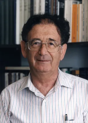 Ein älterer Herr mit Brille und hellem Hemd, der leicht lächelnd in die Kamera blickt; im Hintergrund ist ein gefülltes Bücherregal zu sehen.