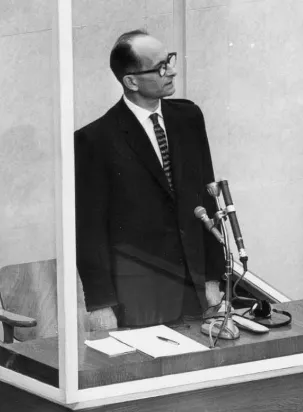 Eichmann trägt Brille, Krawatte und Anzug. Er steht während der Verhandlung in der Glaskabine. Vor ihm drei Mikrofone, auf einer Ablage Papier und ein Stift.