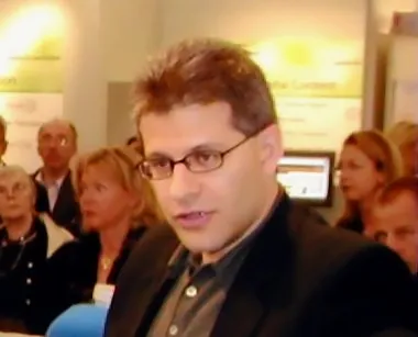 Goldhagen mit kurzem braunem Haar und Brille, er trägt ein Jackett und ein braunes Hemd und ist offenbar in einem öffentlichen Raum in ein Gespräch mit jemand außerhalb des Bildes vertieft.