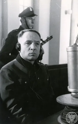 Rudolf Höß bei seiner Aussage, er träg eine hochgeschlossene Jacke und hat einen Kopfhörer. Hinter ihm ein bewaffneter Soldat. Rechts vorne ein Mikrofon.