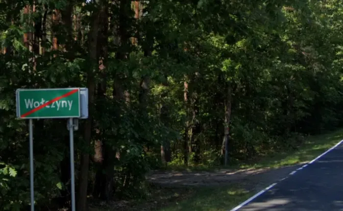Das rechteckige Ortschild links neben der Straße ist grün, die Schrift ist weiß. Der Ortsname Wolczyny ist mit einem roten Strich schräg durchgestrichen: Ortsausgang.