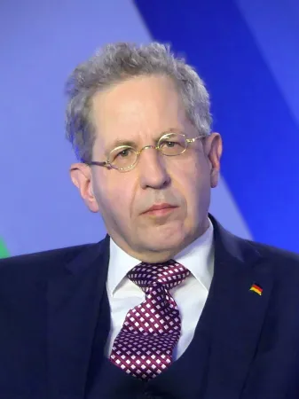 Der Mann mit graumeliertem, zurückgekämmtem Haar trägt eine Nickelbrille, dazu einen Anzug mit violett-weiß gepunkteter Krawatte. Im Revers ein kleines Deutschlandfähnchen. Er schaut den Betrachter etwas missmutig an.
