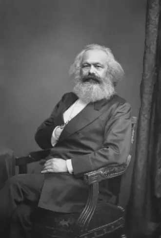 Portrait von Karl Marx mit Vollbart. Er ist im Anzug auf einem Stuhl und hat die rechte Hand unter das Revers geschoben.