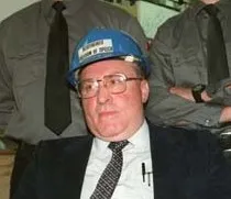 Ernst Zündel mit blauem Schutzhelm und Brille im Anzug, er trägt darunter ein weißes Hemd und eine dunkle Krawatte.