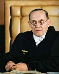 Richter Rothaug auf dem Richterstuhl, er trägt eine Brille und die Richterrobe, vor ihm liegen Akten auf dem Tisch.