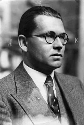 Ein alltäglich aussehender Bürokrat mit beinahe noch jugendlichen Gesichtszügen, glatt zurückgekämmten Haaren, Brille, Anzugjacke, Oberhemd und Krawatte.